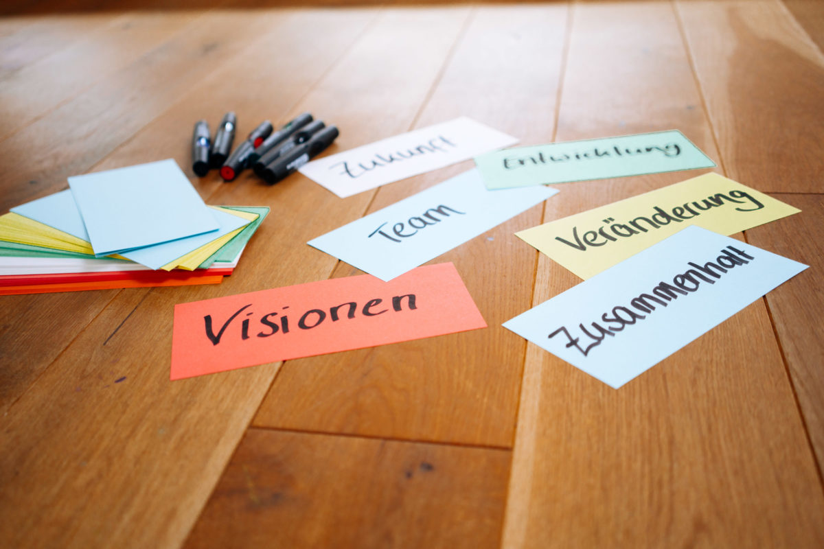 Verschiedenfarbige Moderationskarten mit verschiedenen Wörtern beschriftet liegen zusammen mit Stiften auf einem Parkettboden